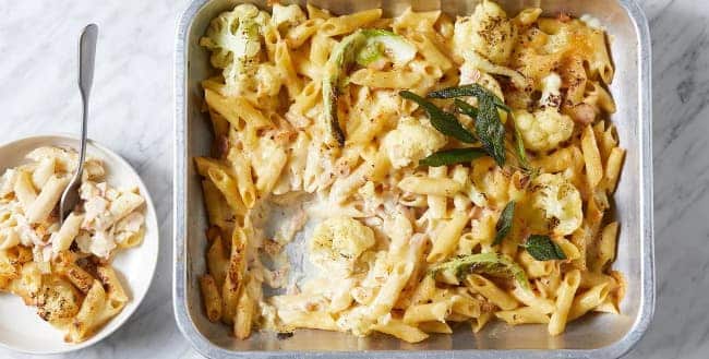 Penne baked with cauliflower and sage in a baking dish with a plate of pasta with a spoon next to it.