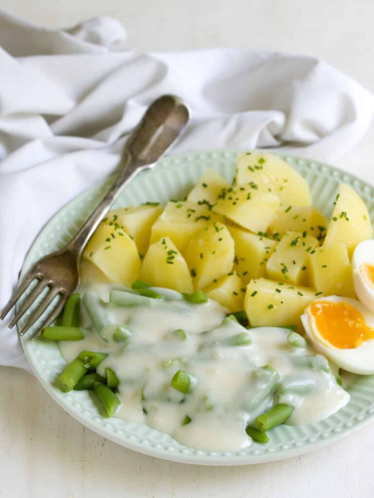 Bílá omáčka s fazolkami servírovaná na talíři s vařenými bramborami, vejcem a vidličkou.