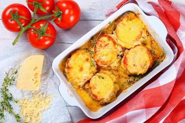 Baked meat with cream sauce and cheese in a baking dish. Fresh tomatoes and a piece of cheese are placed next to it.