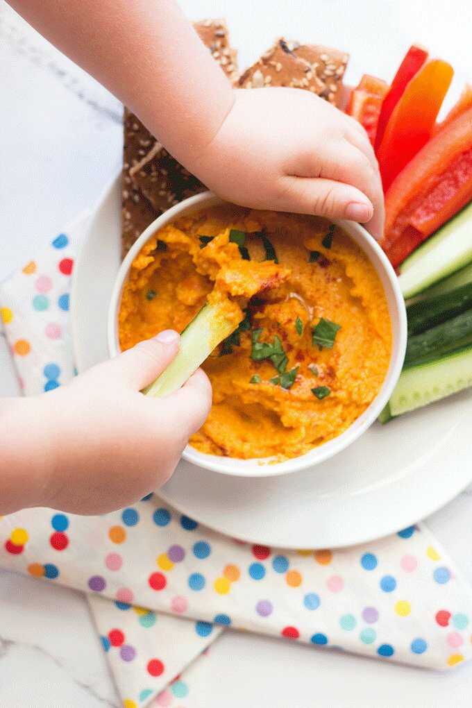 Hände schöpfen Karotten-Hummus aus einer Schüssel. Es wird mit frischem Gemüse und Crackern serviert.