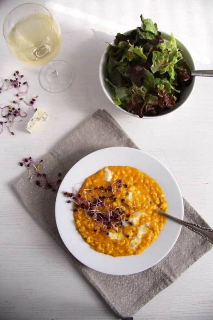 Risotto with gorgonzola and pumpkin in a deep plate with a spoon, decorated with sprouts. Next to it is a bowl of fresh salad and a glass of white wine.