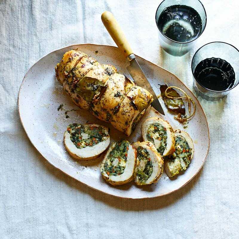 A turkey roll filled with a mixture of rice, spinach, carrots and feta sliced and served on a large plate with a knife and two glasses of water next to it.