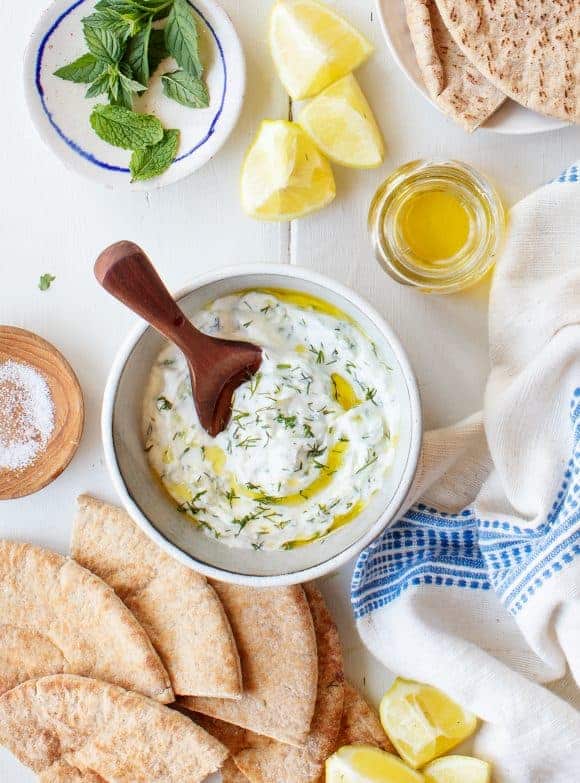 Yogurt dip with cucumber and dill in a bowl, drizzled with olive oil and served with a wooden spoon. Pita bread, a glass of oil, a lemon wedge and a plate with fresh herbs are placed next to it.