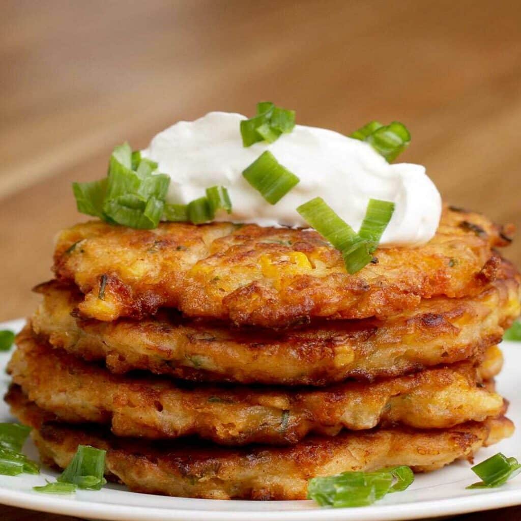 Corn and cheddar pancakes stacked on top of each other, served on a plate and garnished with sour cream and chives.