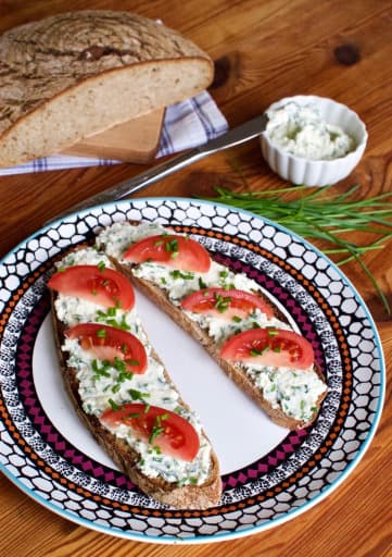 Sliced roll spread with cottage cheese spread and decorated with chives and tomatoes. All this served on a plate, with fresh bread and chives on the side.