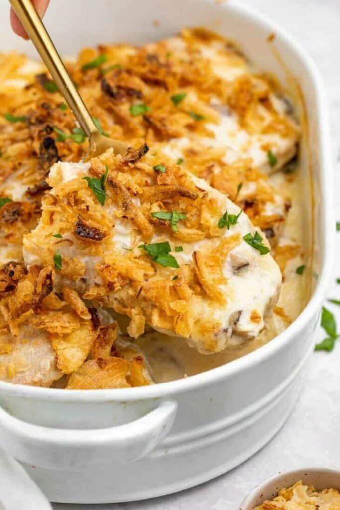 Pork with cheese mixture, fried onions and herbs in a baking dish, scooped with a large ladle.