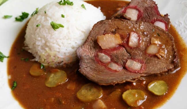 Sauce with pickles, bacon and a slice of beef served on a plate with a scoop of rice.