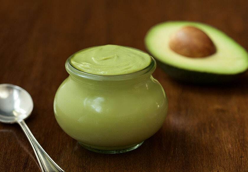 Avocado mayonnaise in a jar with a spoon on one side and half an avocado on the other.