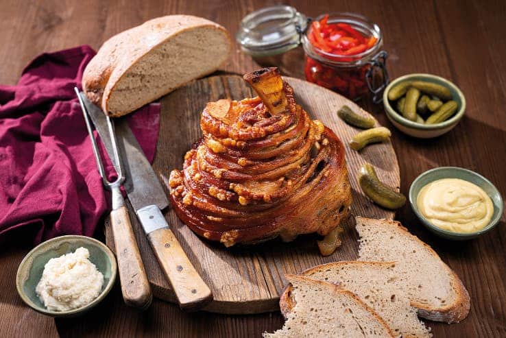 Schweinshaxe auf einem Brett, serviert mit Brot, Essiggurken, Senf und Meerrettich.