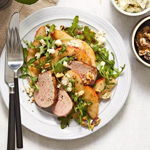 Arugula and pear salad served on a plate with cutlery along with slices of pork tenderloin.