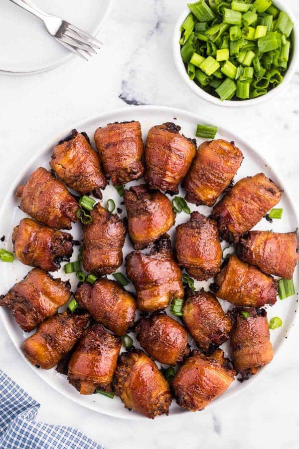 Meat rolls wrapped in bacon, served on a large plate. A bowl with chopped spring onions is placed next to it.