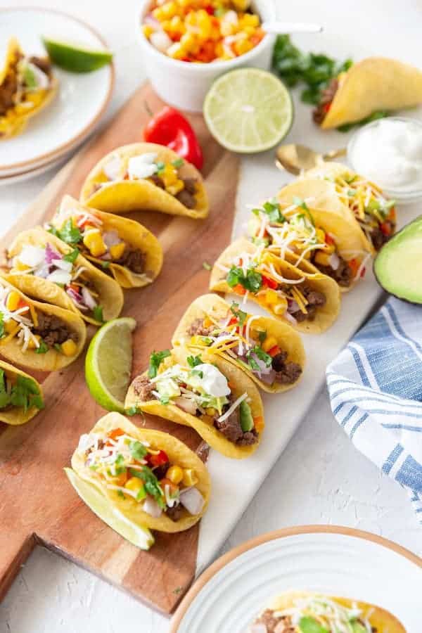 Small tacos with ground beef, salsa, sour cream, cheese and herbs on a cutting board.