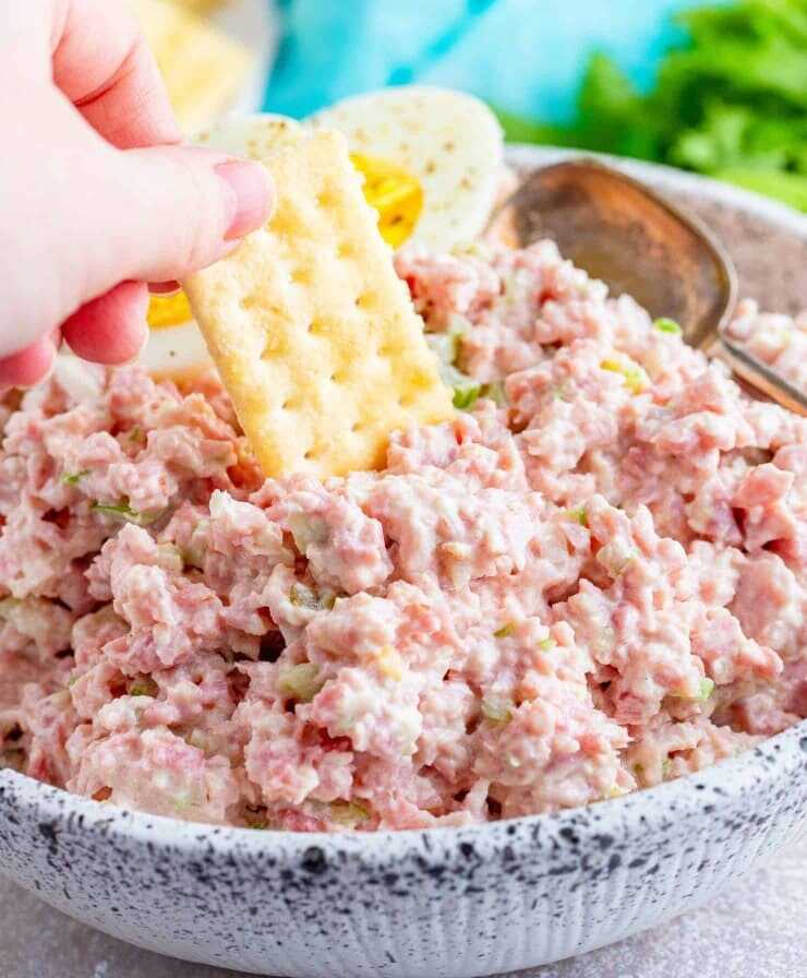 A salad of ham, mayonnaise, celery stalks and onions in a large bowl, into which a hand dips a cracker.