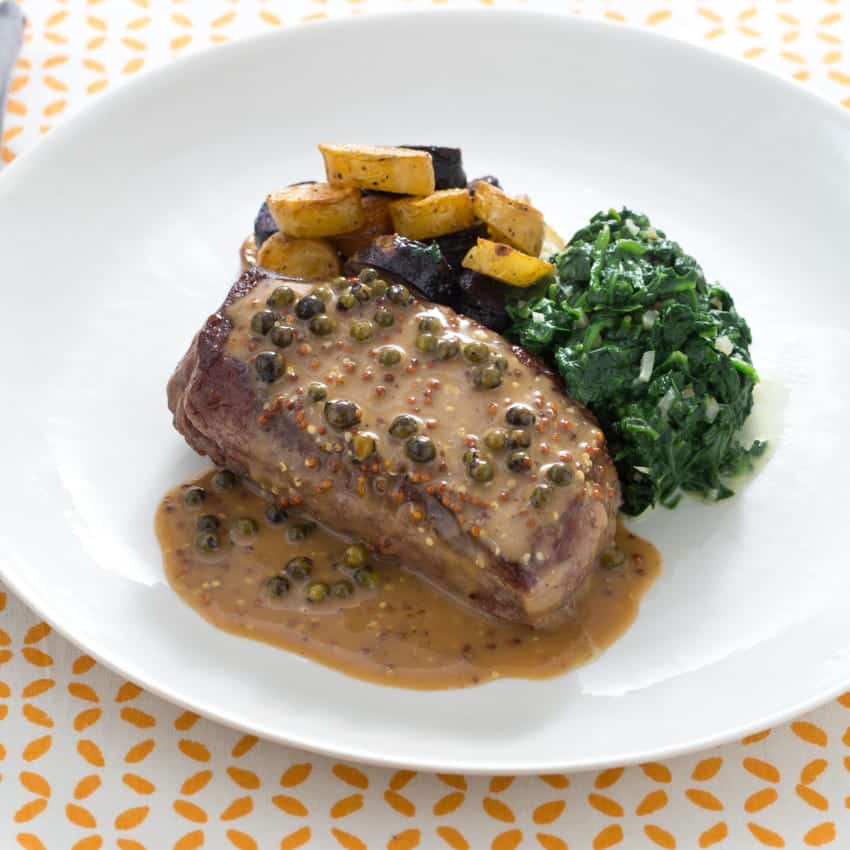 Hovězí steak s krémovou omáčkou, zeleným pepřem, bramborami a špenátem na talíři.