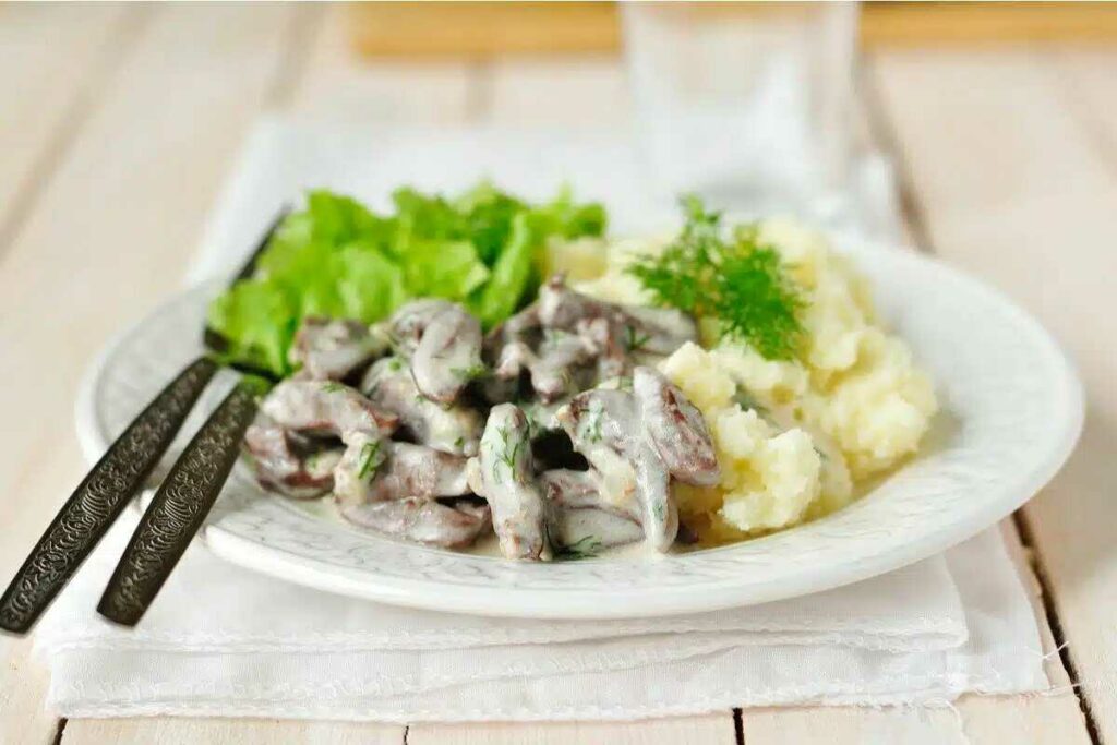 Smetanová játrová omáčka s bramborovou kaší servírovaná na talíři se salátem a s příborem.