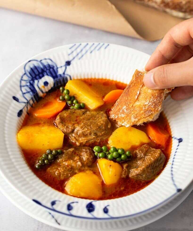 A slightly spicy sauce with tender beef cubes, potatoes, carrots and fresh green pepper on a plate and a hand scooping up this sauce with a pastry.