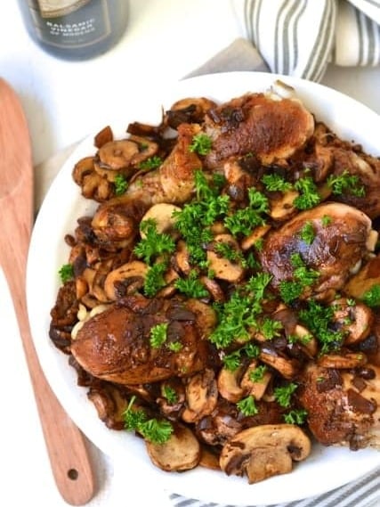 Pilze, Schalotten und Balsamico-Sauce gemischt mit köstlichen Hähnchenkeulen, garniert mit frischen Kräutern und serviert auf einem Teller.