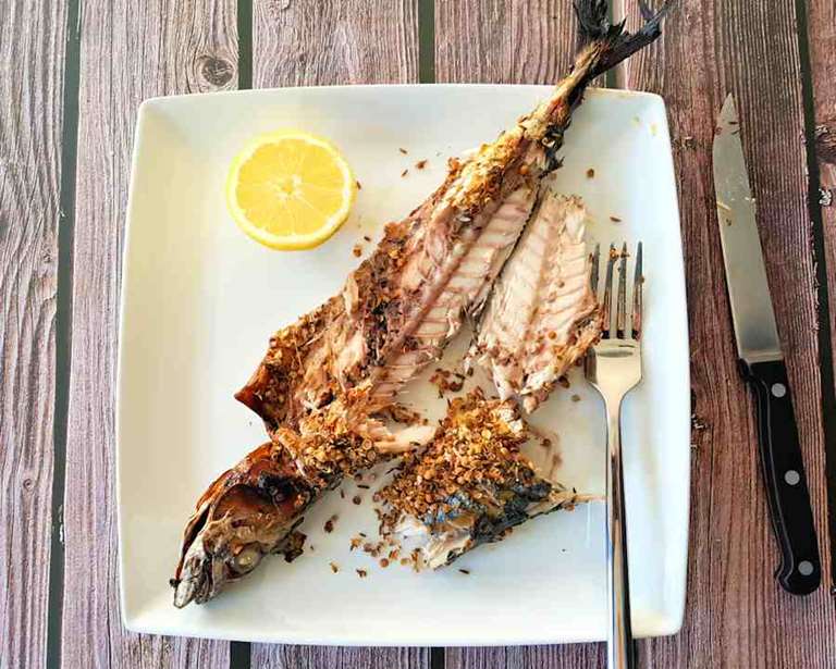 Gegrillte ganze Makrele mit Knoblauch-, Kreuzkümmel- und Korianderkruste auf einem Teller mit einer Gabel und einer Zitronenscheibe. Daneben wird ein Messer platziert.