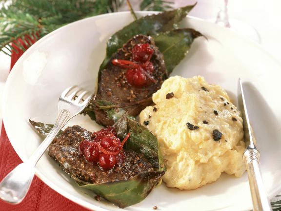 Medailonky ze srnčího servírované s omáčkou z brusinek a červeného vína a podávané s kaší z brambor s lanýži na talíři s příborem.