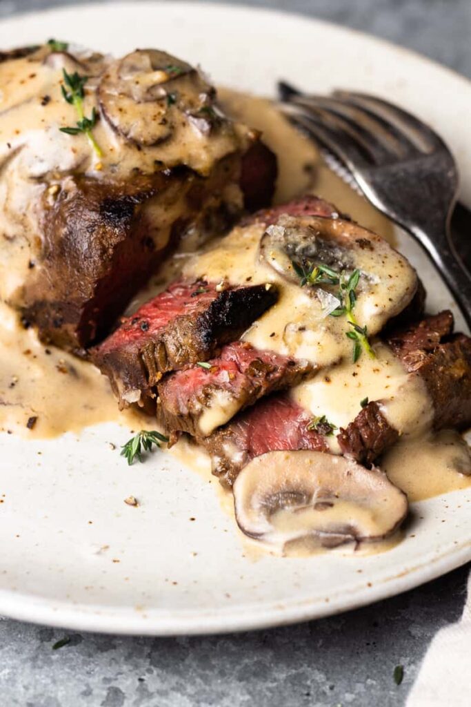 Marinované steaky ze srnčího masa opečené na pánvi a podávané s dokonalou krémovou omáčkou ze žampionů a bílého vína na talíři s příborem.
