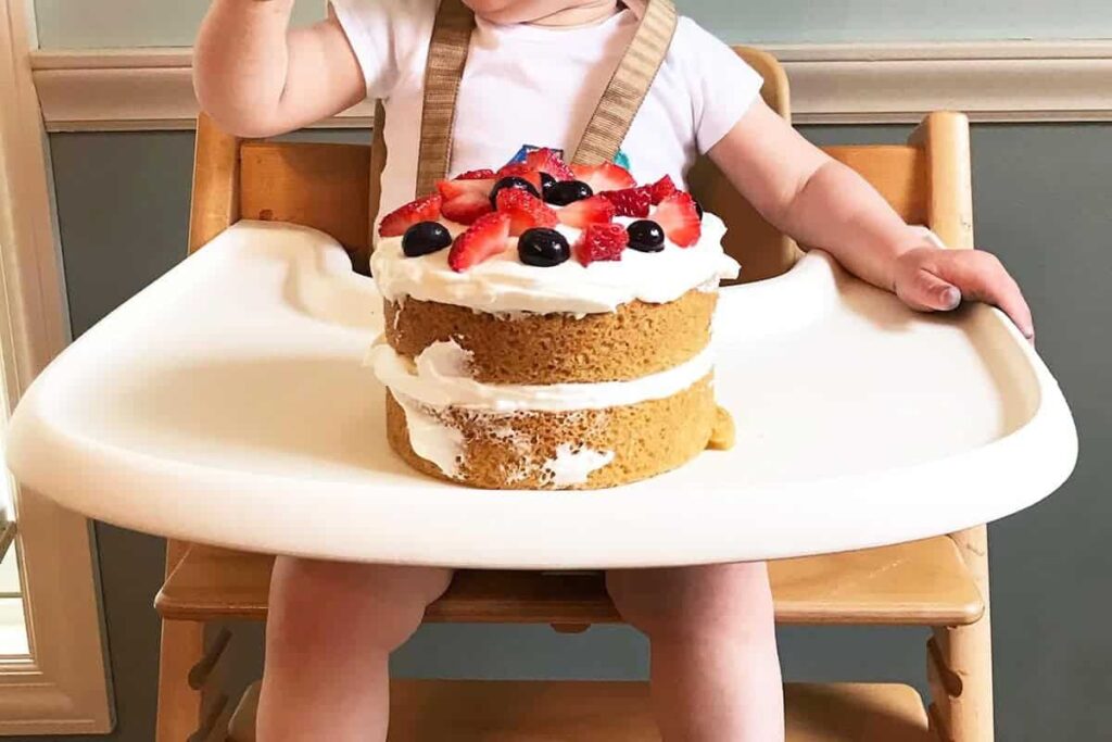 Zdravý dětský dort s krémem ozdobený čerstvým ovocem na stolečku u jídelní židličky, ve které sedí dítě.