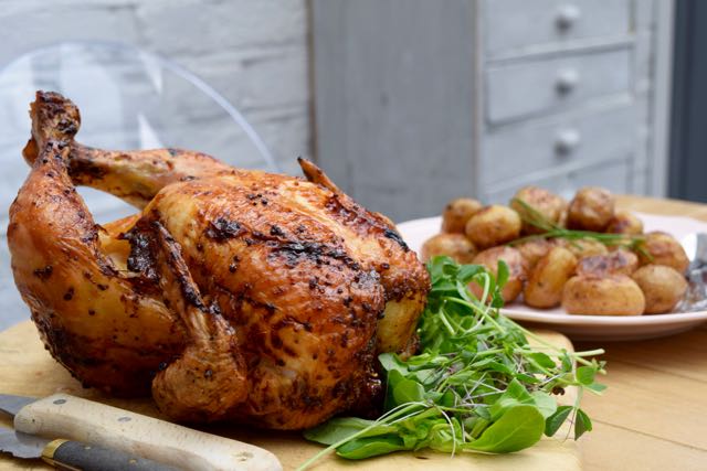 Upečené kuře na prkýnku s vedle položeným talířem s baby bramborami a salátem.